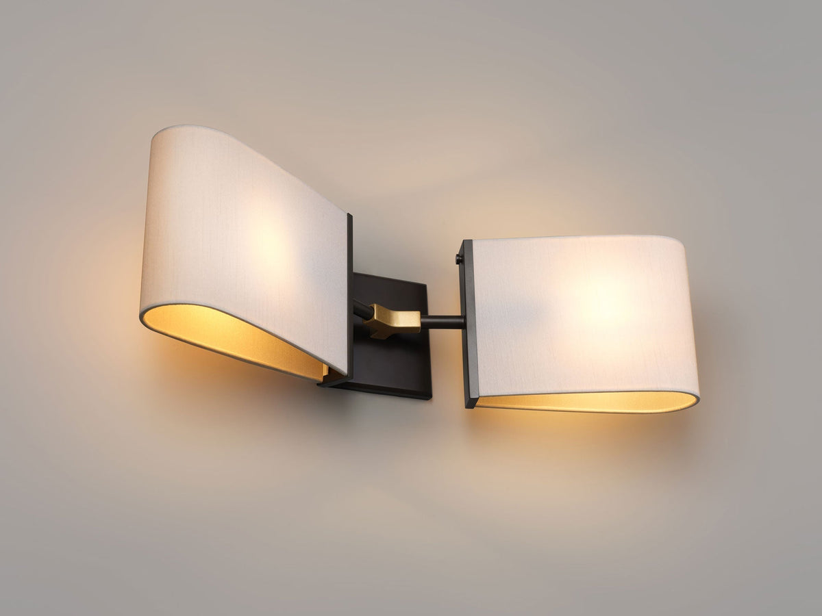 Lit double wall sconce with two ivory fabric shades angled outward, a dark bronze backplate and brass joint on a grey wall